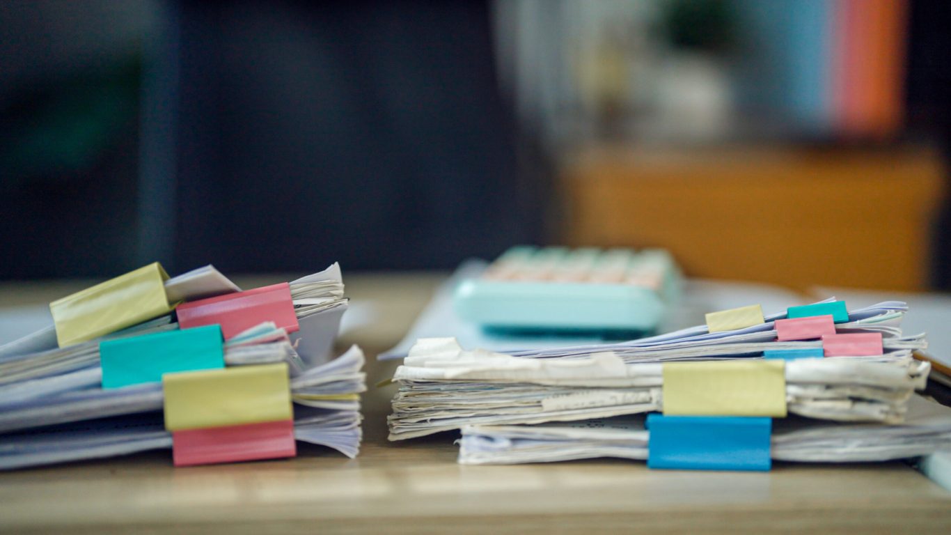 Stacks of papers, colorful tabs, and a calculator sit on a desk. The scene suggests busy work or urgent organization. Focus is on details, not people or emotions. Ideal for business, finance