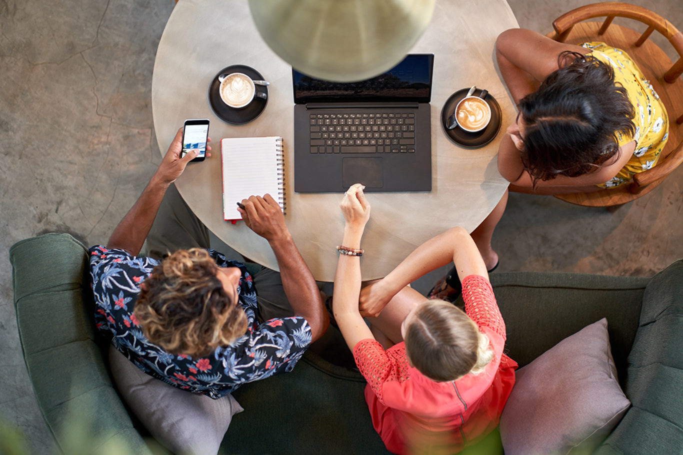 Unposed overhead shot of two mixed race young women and man wearing bright fashionable clothing, working together over fair-trade coffee with a laptop computer at a round table in a trendy hipster cafe and restaurant