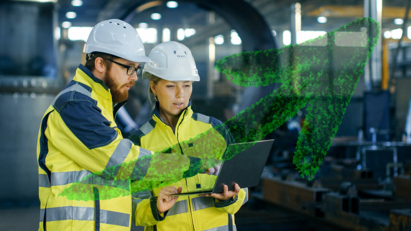 Male and Female Industrial Engineers in Hard Hats Discuss New Project while Using Laptop. They Make Showing Gestures.They Work in a Heavy Industry Manufacturing Factory.