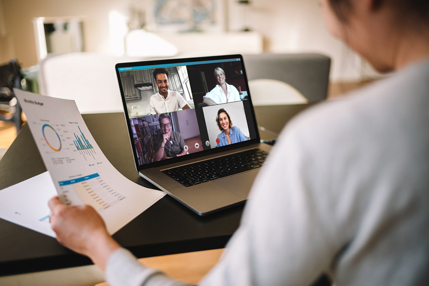 Woman holding a business report working at home having a video conference with colleagues. Over the shoulder view of a businesswoman having online business meeting from home.