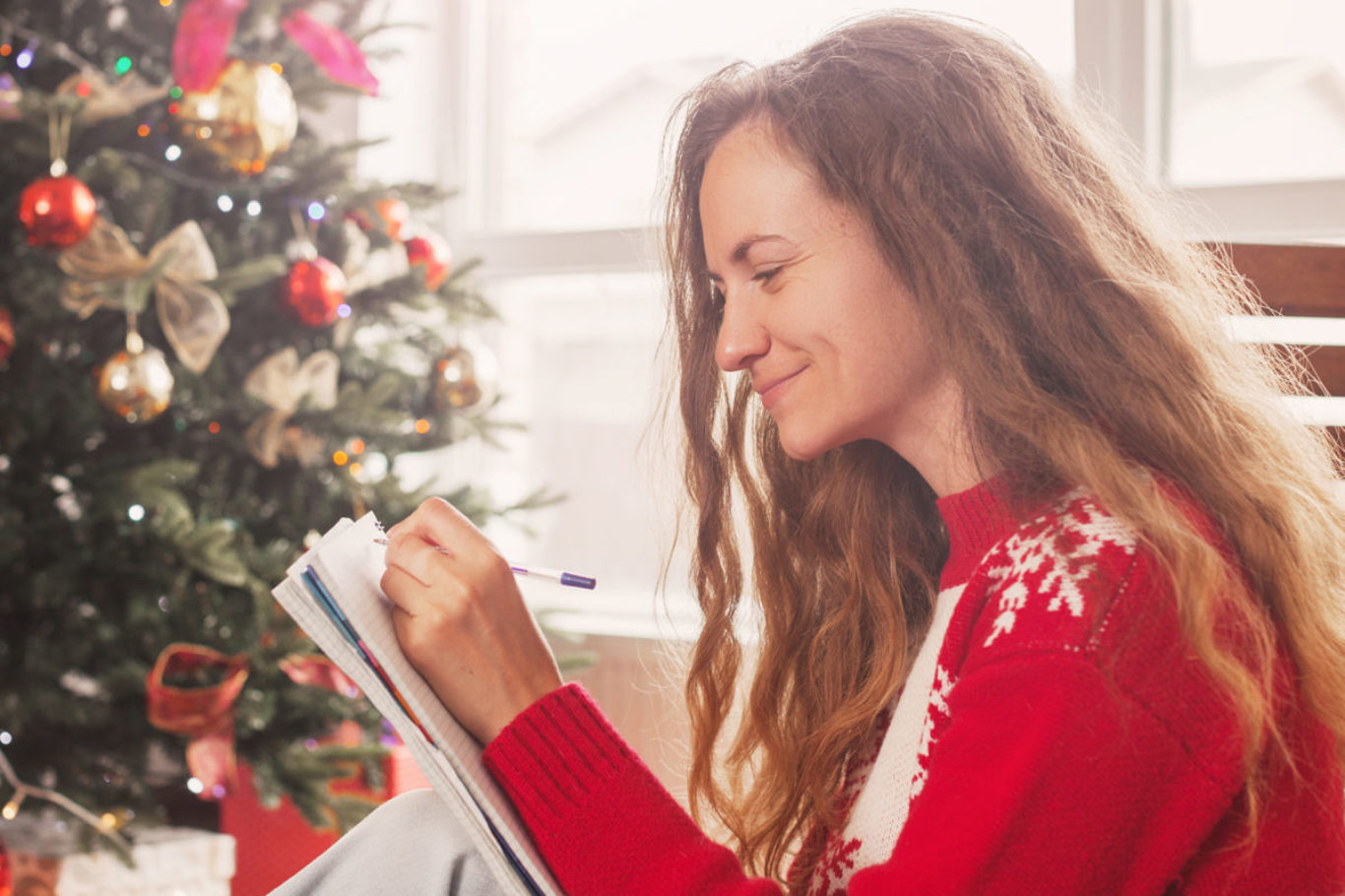 Happy woman writing list of presents for her family members, sitting at home among Christmas wrapped gift boxes. Dreamy woman taking notes in notebook and smiling