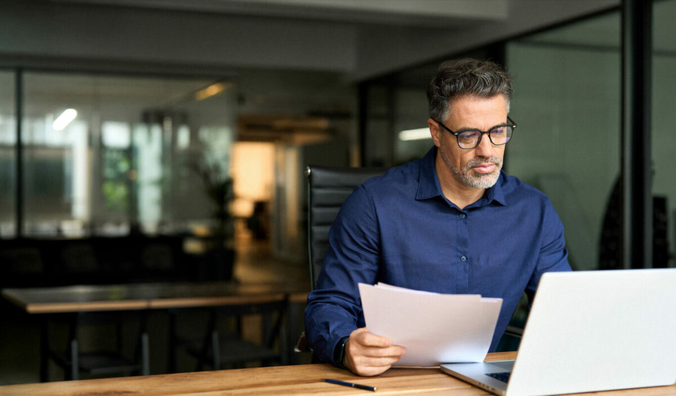 Middle aged professional business man using laptop computer reading document at work. Busy mature male account bank manager executive working on laptop checking financial report in office. Copy space.