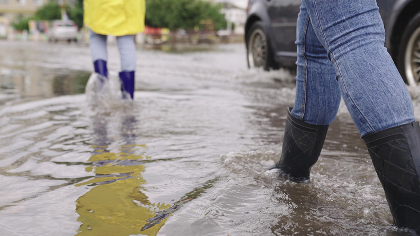 girls in raincoats and rubber boots walk along road flooded with torrential rains, their feet walk through puddles city, splashing water to the sides, the flood is on street, car is driving on water.