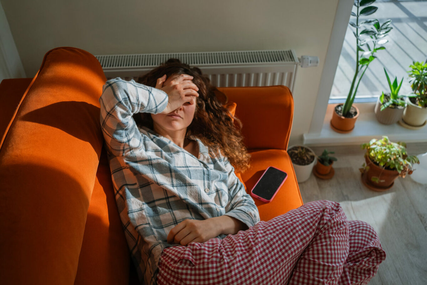 Woman feeling unwell and exhausted while lying on an orange sofa, covering her eyes with a hand, suffering from toxicosis, nausea, or a headache, indicating discomfort and illness