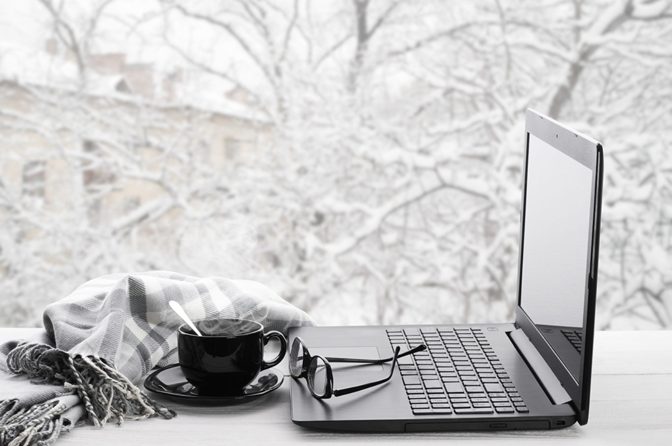 Cozy winter still life: laptop, glasses, cup of hot coffee and warm plaid on windowsill against snow landscape from outside.