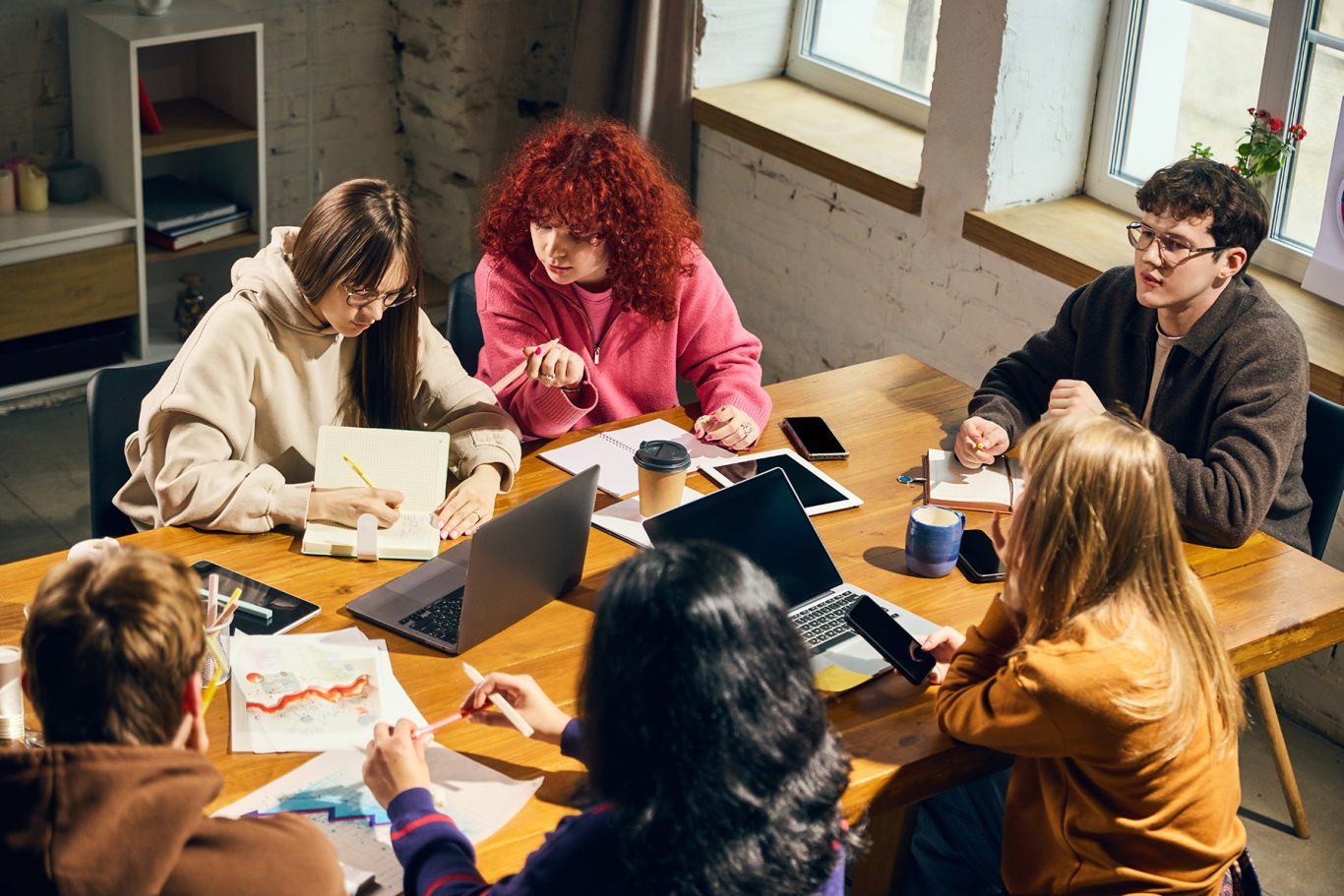 Young people collaborating around wooden table with laptops and charts. Concept of business planning, creative agency work, student workshop, teamwork, analytics, and office training.