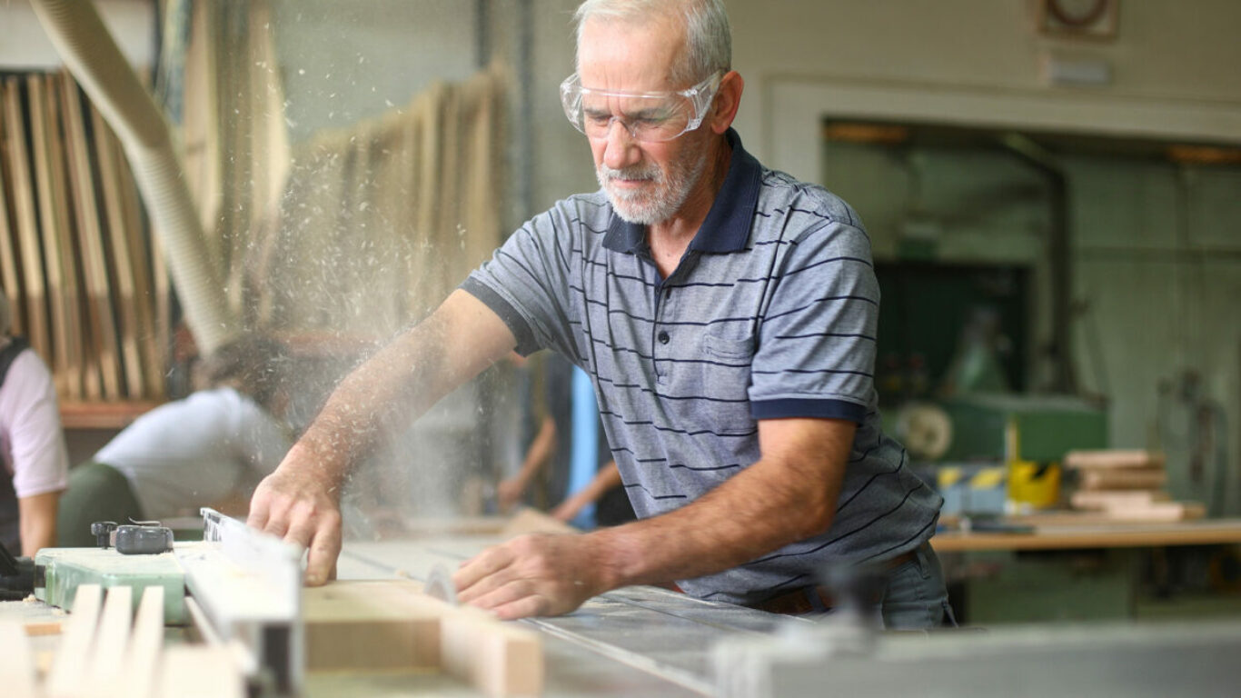 Mature adult carpenter using a table saw, cutting a piece of wood and making sawdust fly in a busy woodworking shop