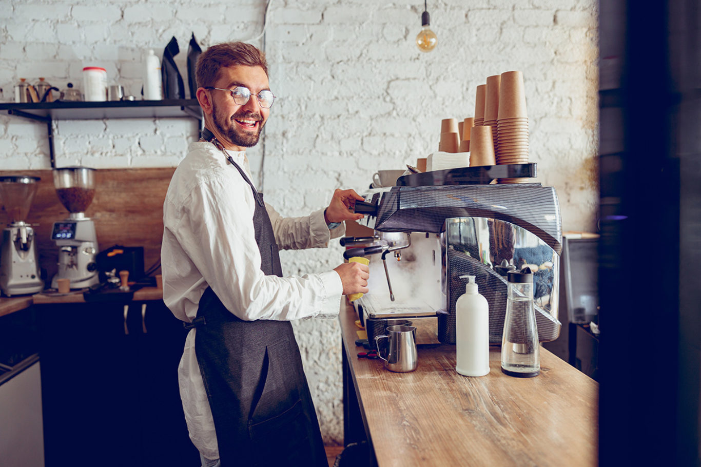Joyful bearded man in apron looking at camera and smiling while using professional espresso machine in coffee shop